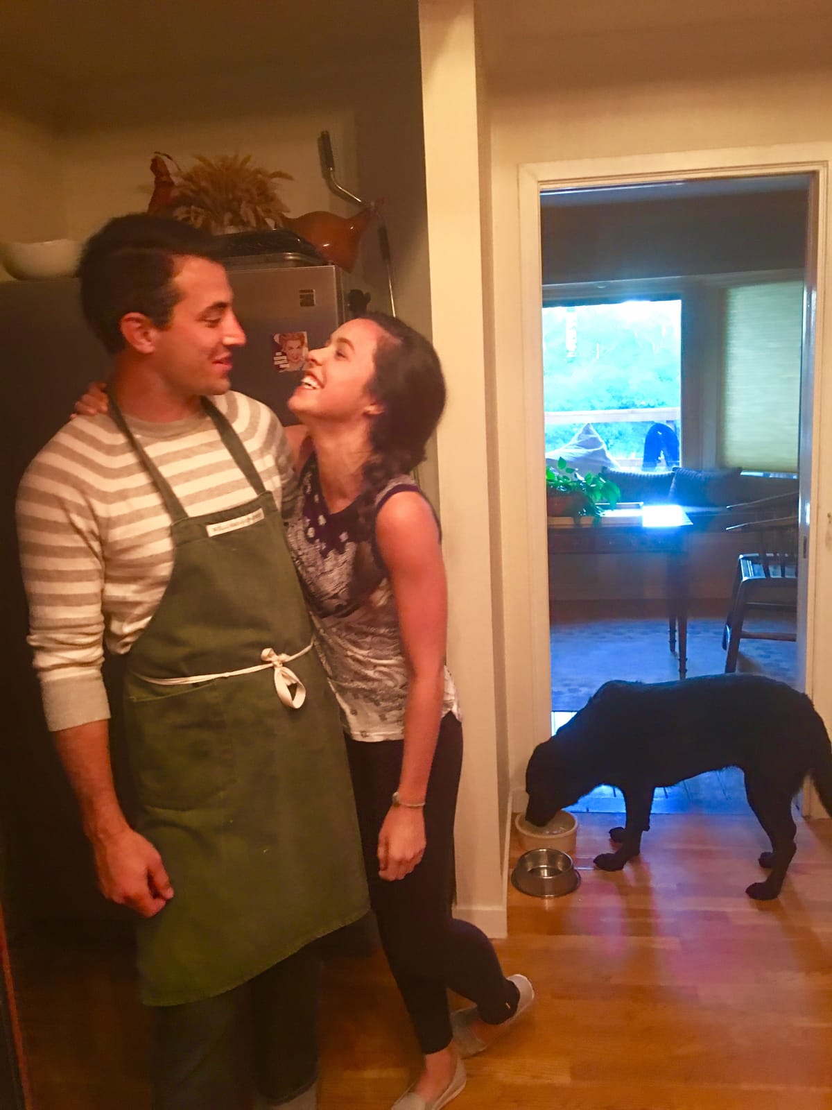 Megan and James laughing in the kitchen while a black lab drinks from a bowl in the next room