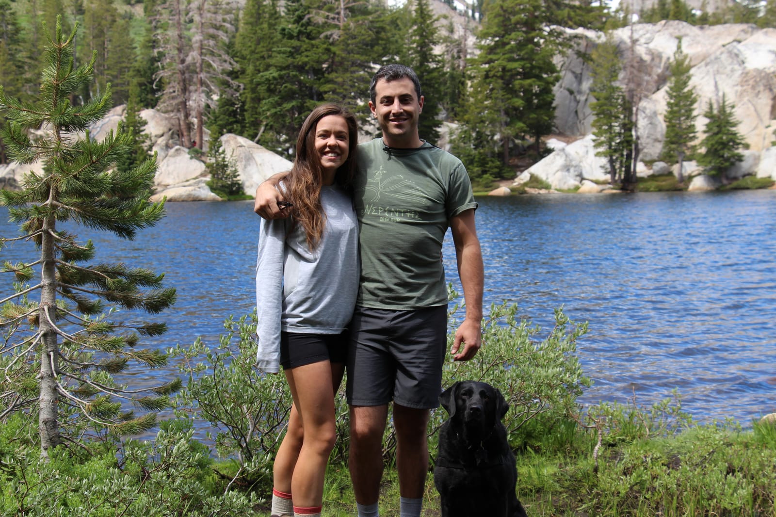 Megan and James with a black lab in front of an alpine lake ringed by granite and pines